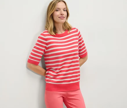 Frau lehnt an Wand, trägt gestreiften Feinstrickpullover und pinke Hose.