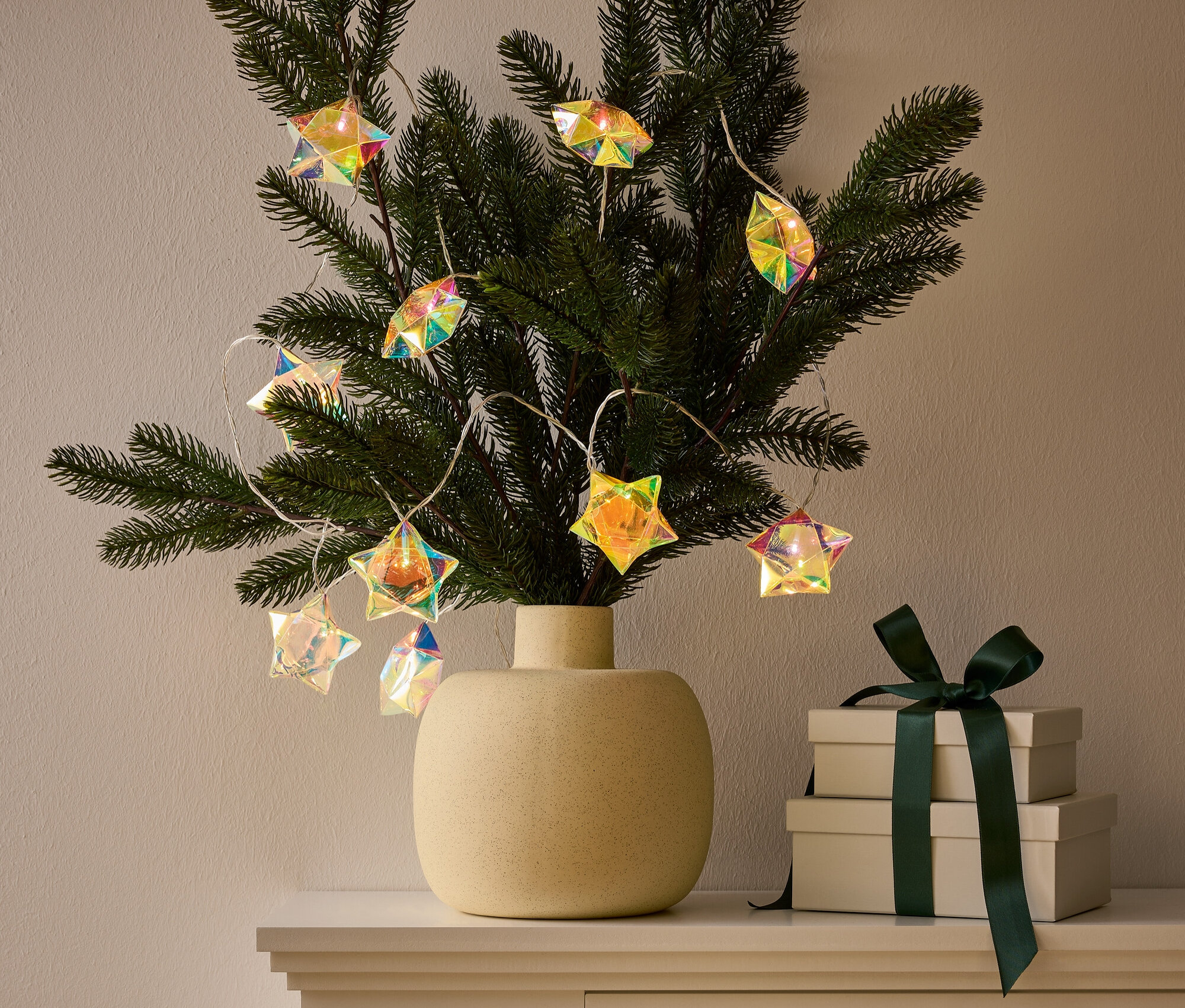 Eine Vase mit einem mit sternförmigen Lichterketten geschmückten Zweig steht auf einem Schrank neben zwei Geschenkboxen mit dunkelgrünem Band.