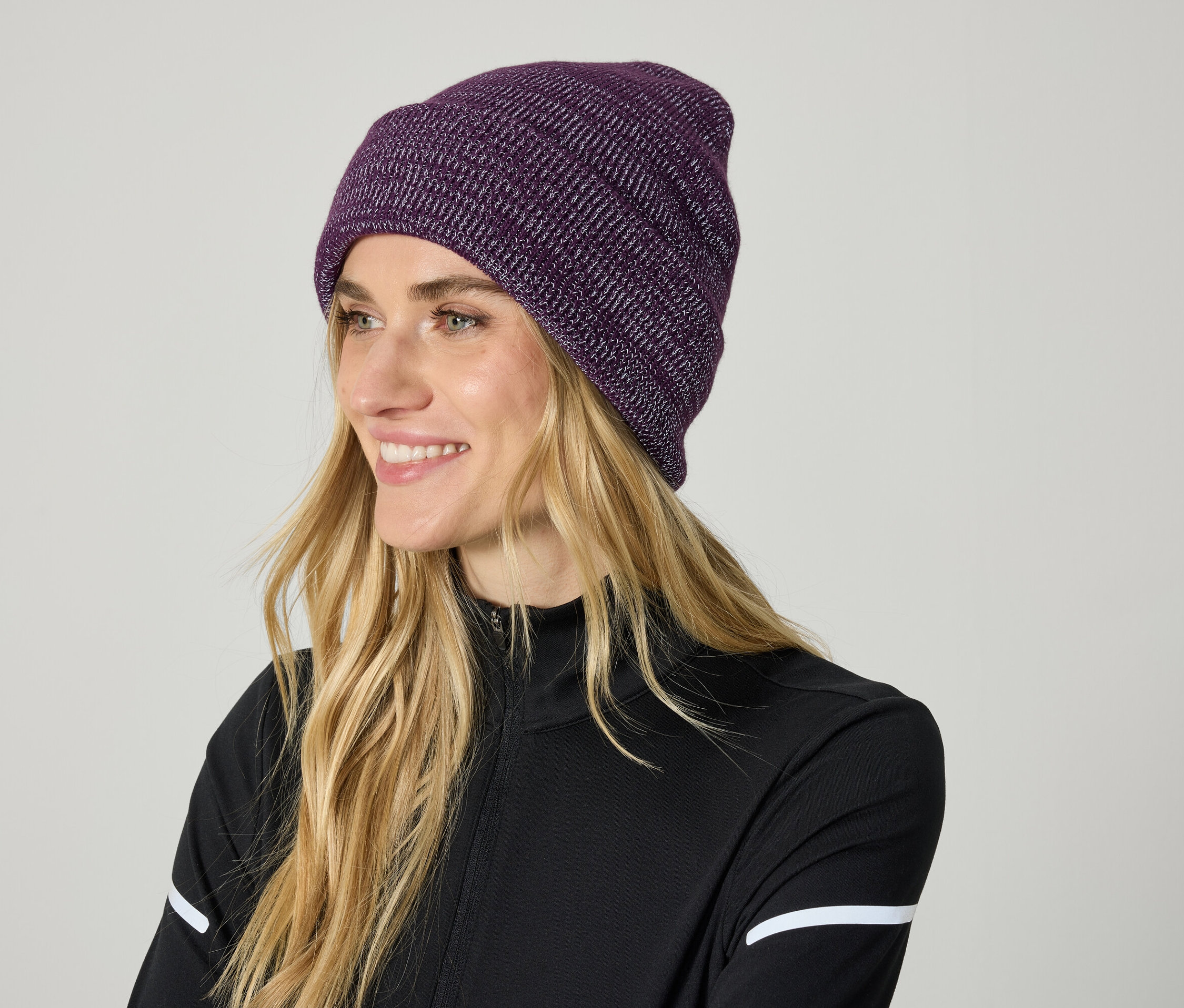 Frau mit blonden Haaren und violetter Strickmütze lächelt.