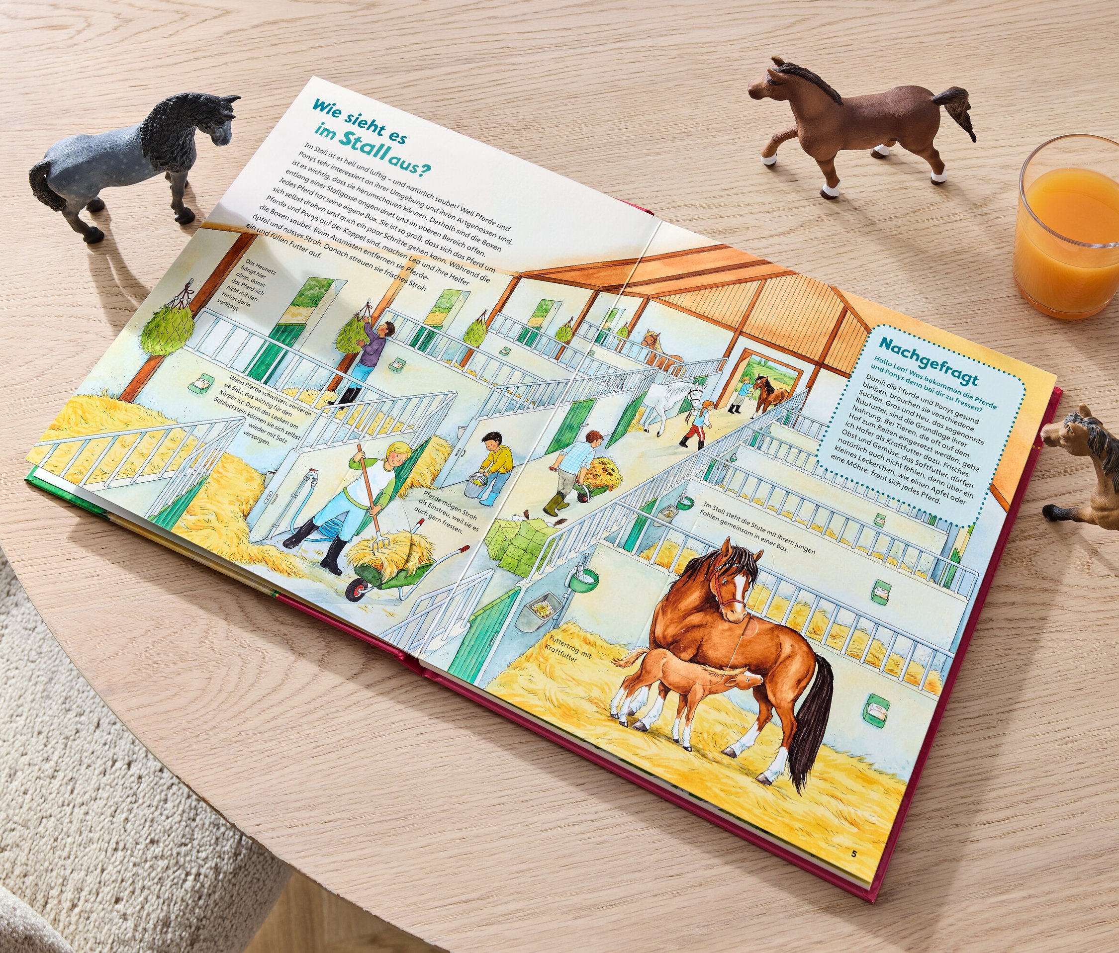 Ein Buch mit einer Illustration von Pferden im Stall liegt auf einem Tisch neben einem Glas Saft und drei Pferdefiguren.