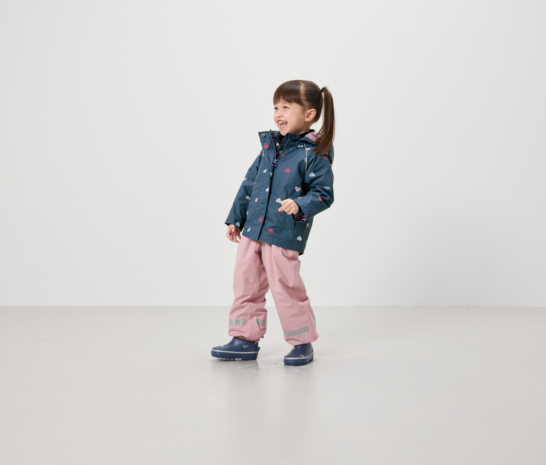 Ein lächelndes Mädchen in einer Jacke mit Herzmuster, rosafarbener Hose und blauen Schuhen steht vor einem hellen Hintergrund.