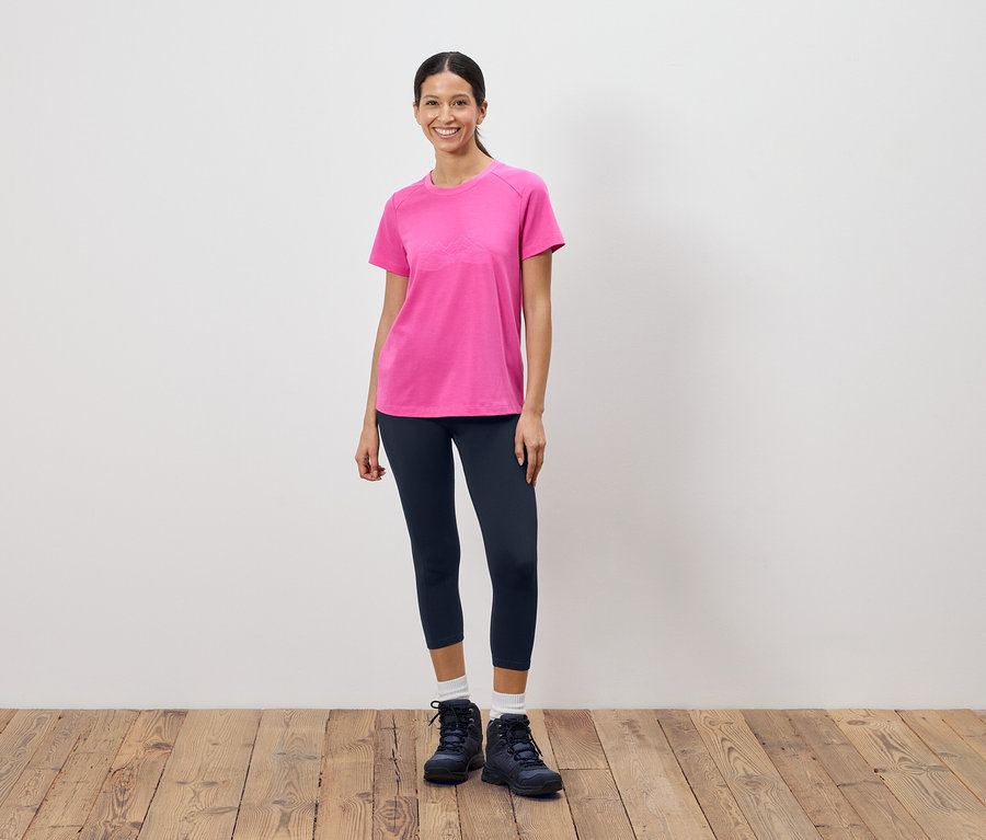 Frau steht vor einer weißen Wand und trägt ein pinkes T-Shirt, dunkle Leggings und Wanderschuhe.