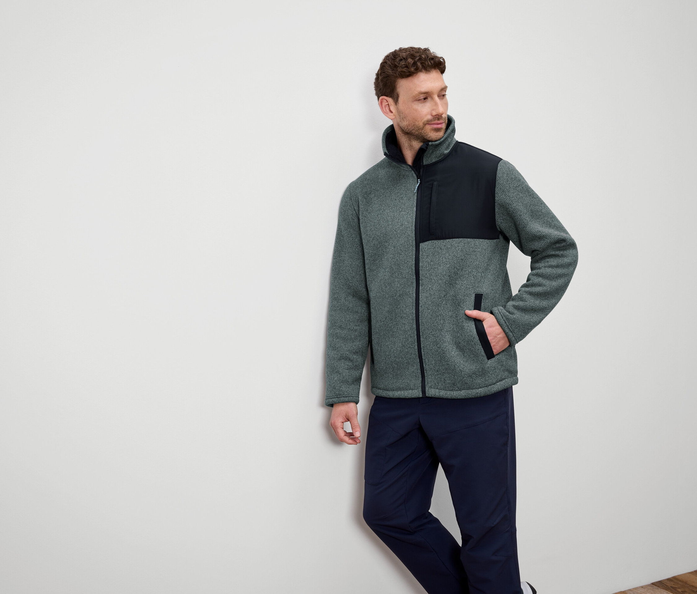 Ein Mann lehnt an einer Wand und trägt eine graue Fleecejacke mit schwarzen Schultern und dunkelblaue Hosen.