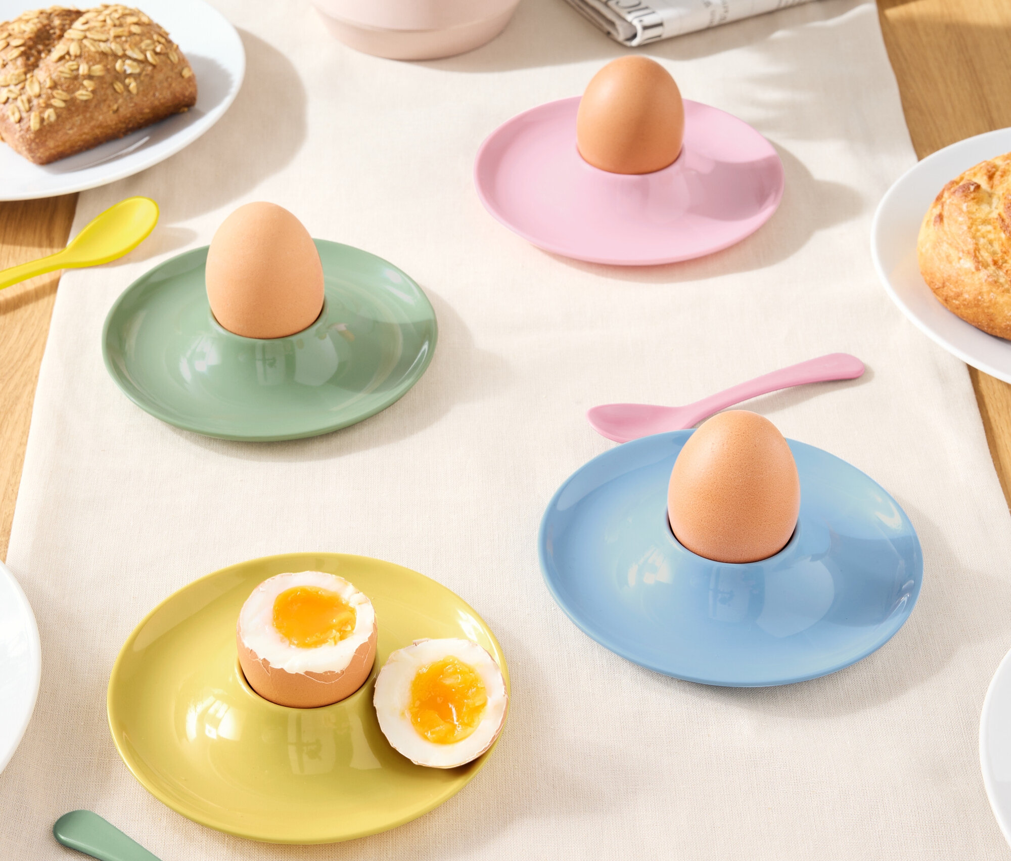 Verschiedenfarbige Eierbecher mit Eiern und Löffeln auf einer Tischdecke.