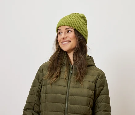 Lächelnde Frau trägt eine olivgrüne Steppjacke und eine Strickmütze.