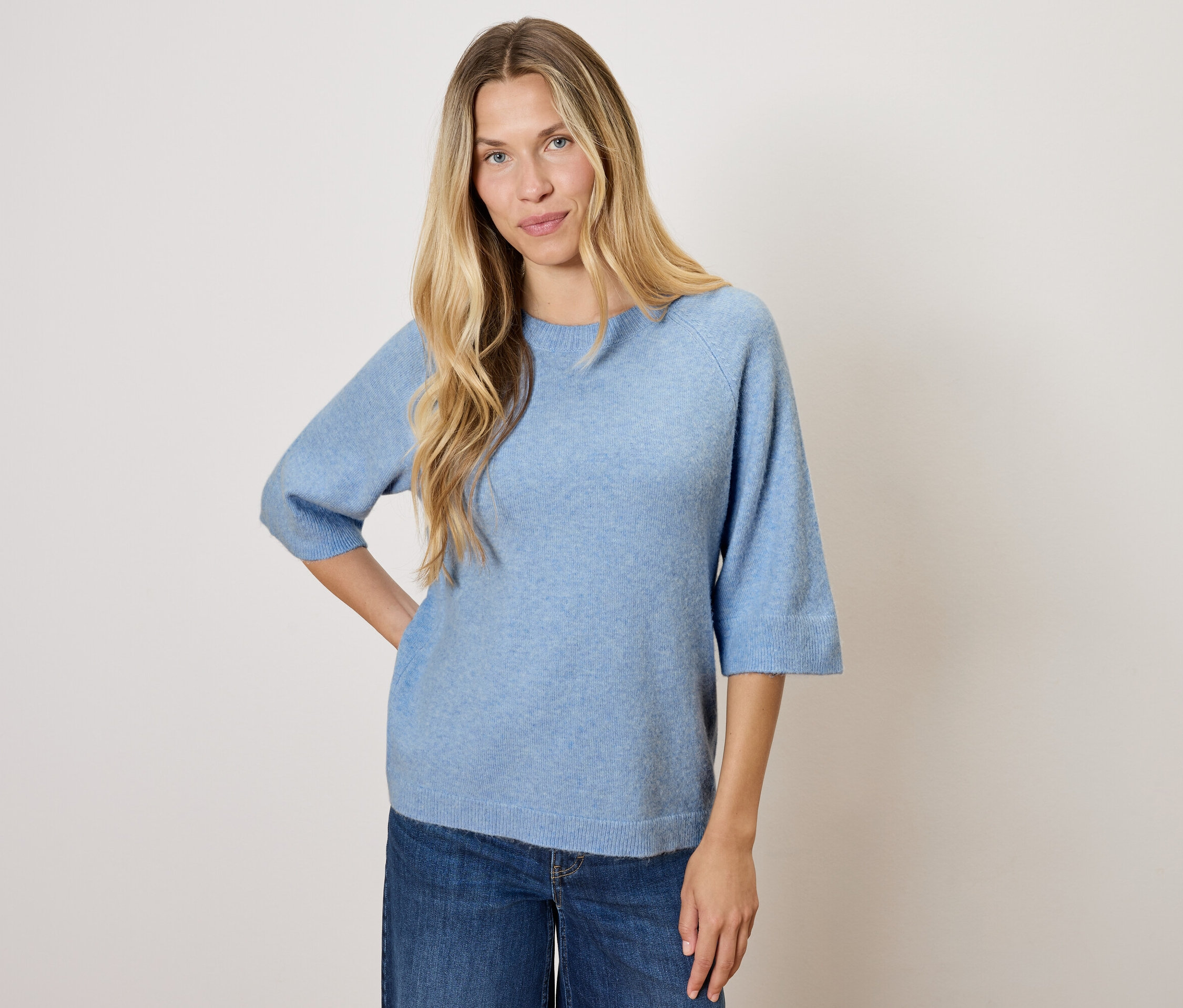 Frau mit blonden Haaren posiert in einem blauen Pullover und Jeans.