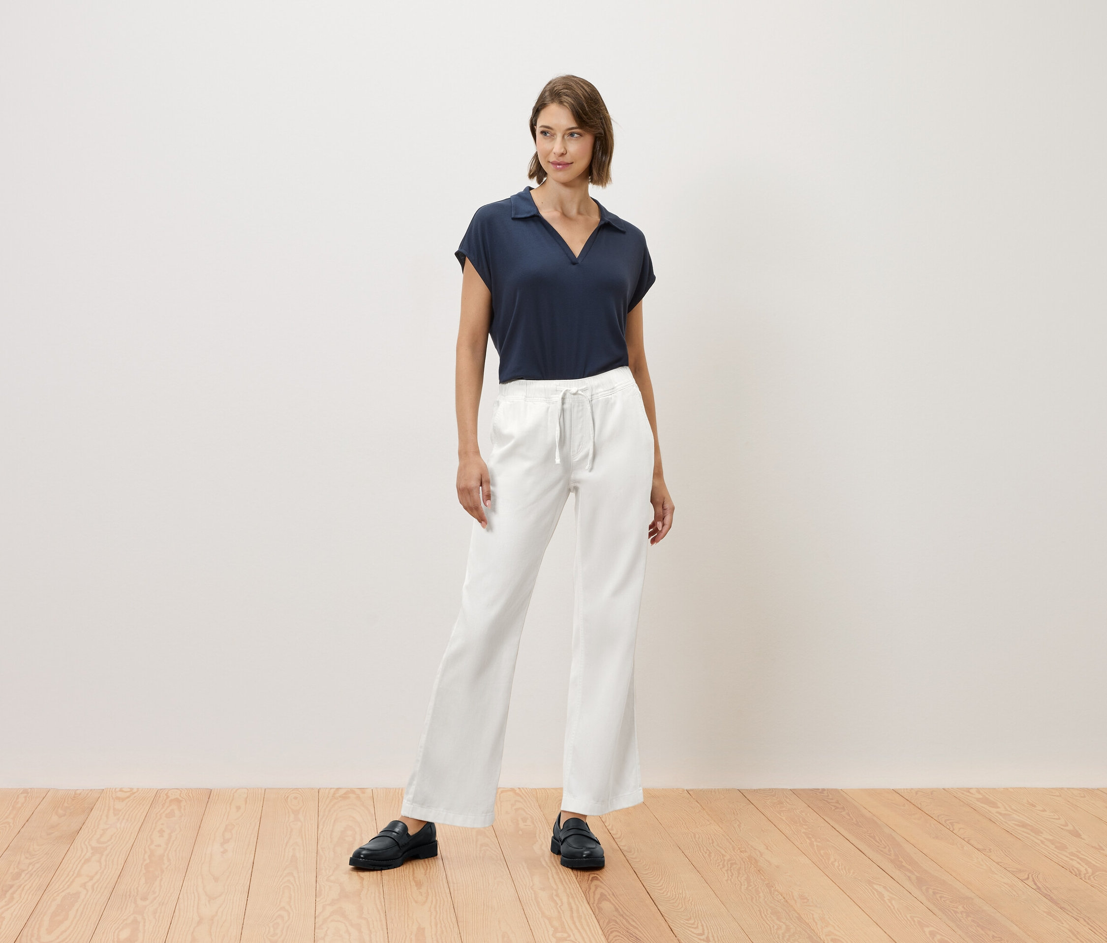 Eine Frau steht auf einem Holzboden und trägt ein marineblaues T-Shirt und eine weiße Hose.