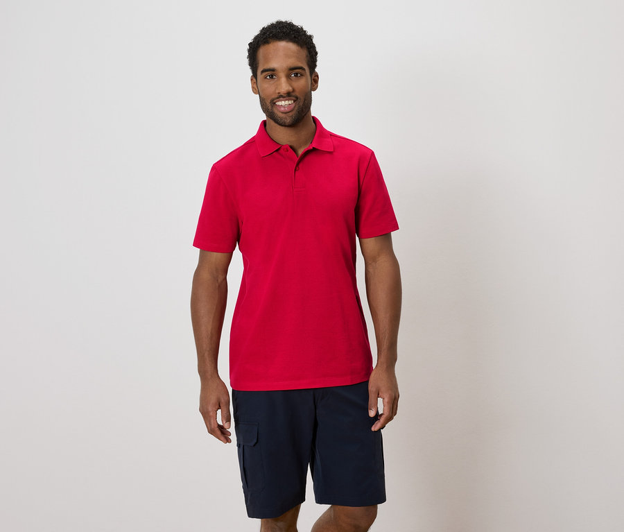 Mann mit rotem Poloshirt und blauen Shorts steht vor einer weißen Wand.
