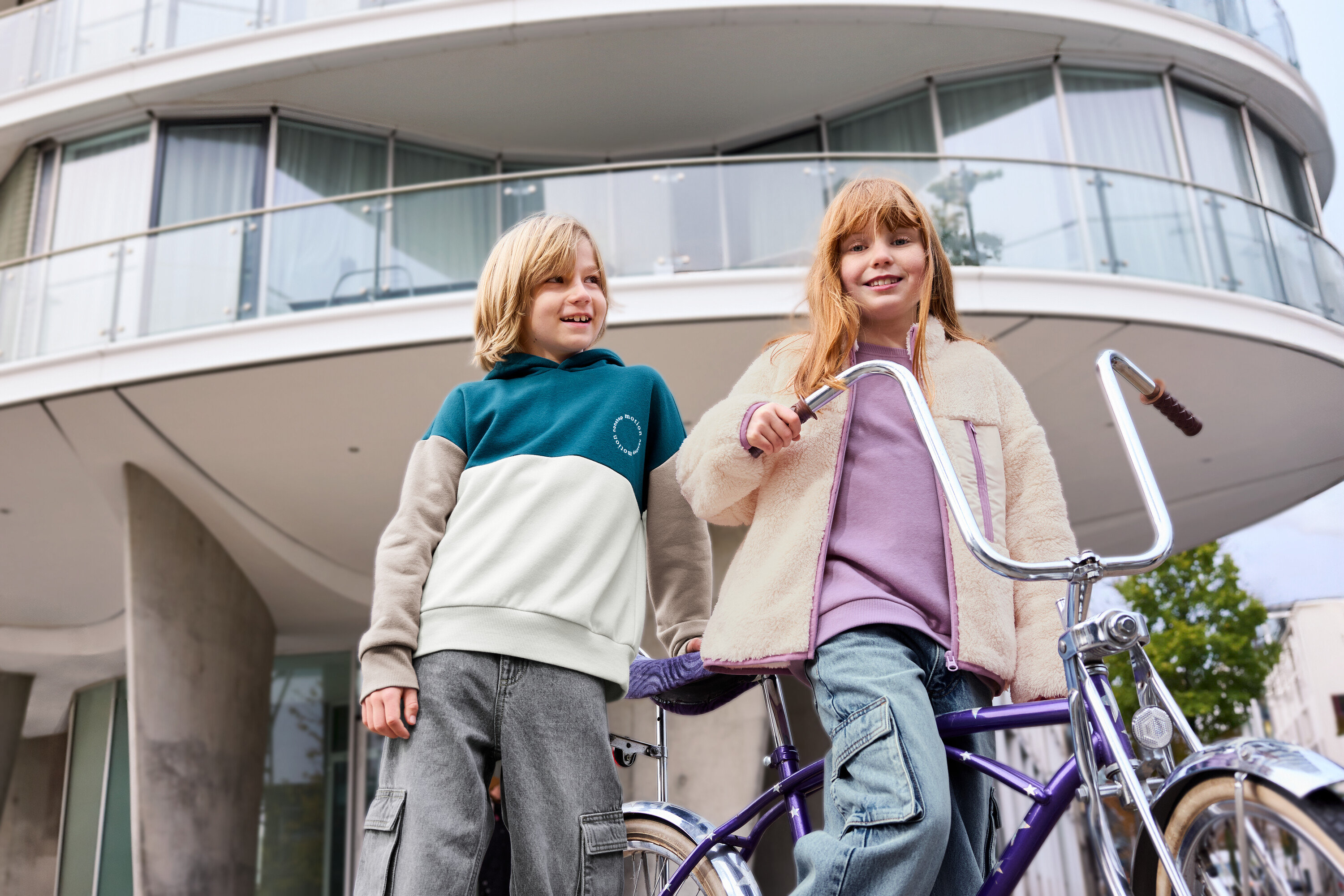 Zwei Kinder stehen vor einem Gebäude, ein Kind trägt ein lavendelfarbenes Kinder-Sweatshirt mit angerauter Innenseite.