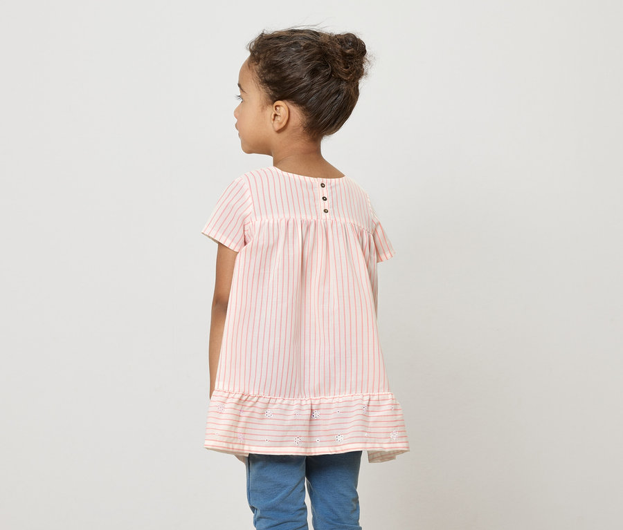 Ein junges Mädchen steht mit dem Rücken zur Kamera und trägt ein rosa-weiß gestreiftes Kleid und blaue Jeans.