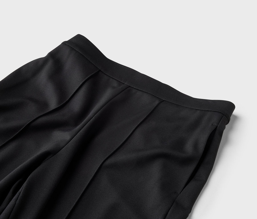 Nahaufnahme der Taille einer schwarzen Hose mit Bundfalten und einer Seitentasche vor grauem Hintergrund.