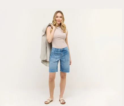Frau mit beigem Oberteil und blauen Angels Denim »Lia Urban« Shorts. Sie hat einen Blazer über der Schulter.