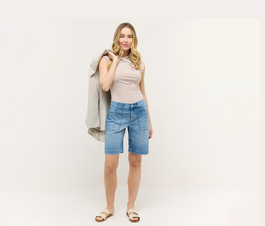 Frau mit beigem Oberteil und blauen Angels Denim »Lia Urban« Shorts. Sie hat einen Blazer über der Schulter.