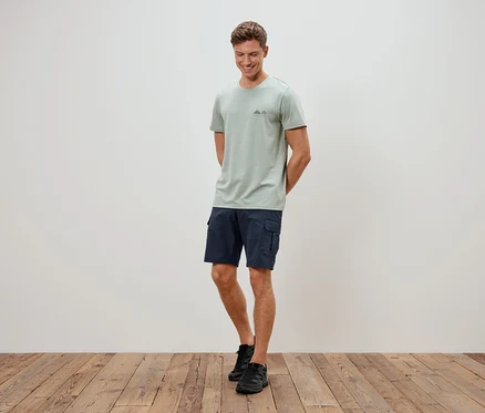 Mann steht vor einer Wand, trägt ein hellgrünes T-Shirt, blaue Shorts und schwarze Schuhe.