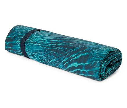 Eine aufgerollte türkisfarbene Strandmatte mit einem Muster, das an Wasserkräuselungen erinnert, wird von einem marineblauen Gummiband zusammengehalten.