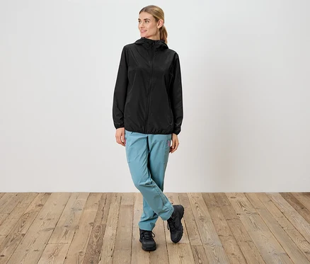 Frau in schwarzer Regenjacke mit Kapuze, hellblauen Hosen und schwarzen Schuhen steht vor einer weißen Wand.