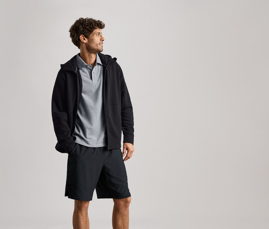 Mann trägt eine schwarze Sweatjacke über einem grauen Polohemd und schwarzen Shorts.