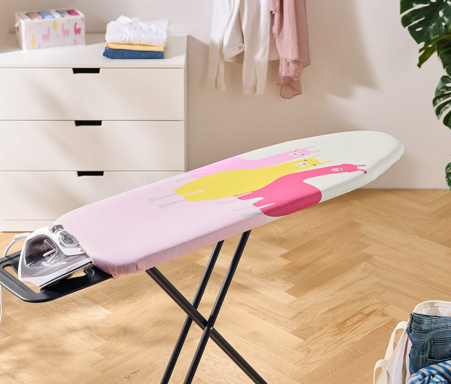 Ein Bügeleisen steht auf einem Bügelbrett mit einem rosa Bezug mit gelben und pinken Lamas. Im Hintergrund befinden sich eine Kommode, Wäsche und eine Zimmerpflanze.