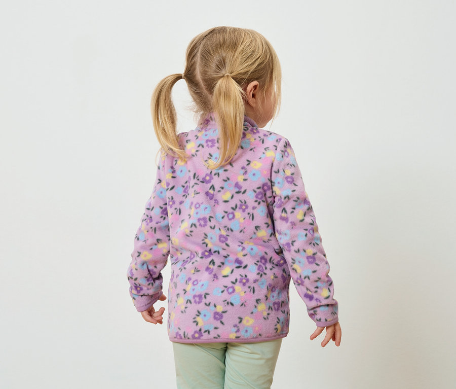 Rückansicht eines kleinen Mädchens mit blonden Zöpfen, das eine lilafarbene Fleecejacke mit Blumenmuster und grüne Hosen trägt.
