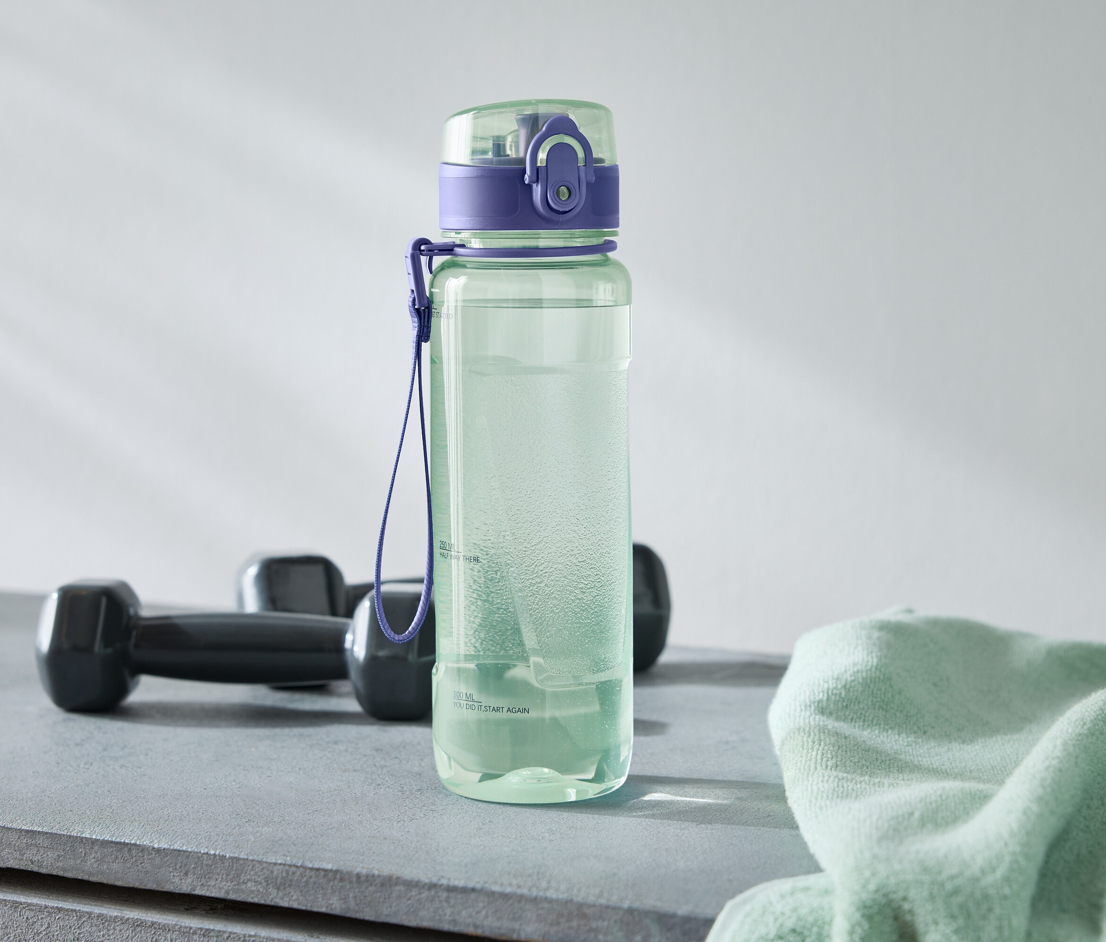 Eine grüne Wasserflasche mit lila Deckel steht neben zwei schwarzen Hanteln und einem hellgrünen Handtuch auf einem grauen Untergrund.