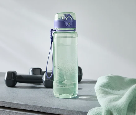 Eine grüne Wasserflasche mit lila Deckel steht neben zwei schwarzen Hanteln und einem hellgrünen Handtuch auf einem grauen Untergrund.