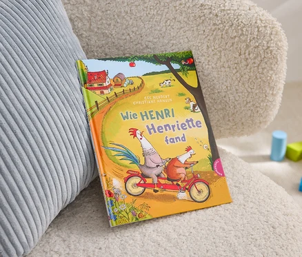 Das Kinderbuch "Wie Henri Henriette fand" liegt auf einem Sessel neben einem grauen Kissen und Bauklötzen.