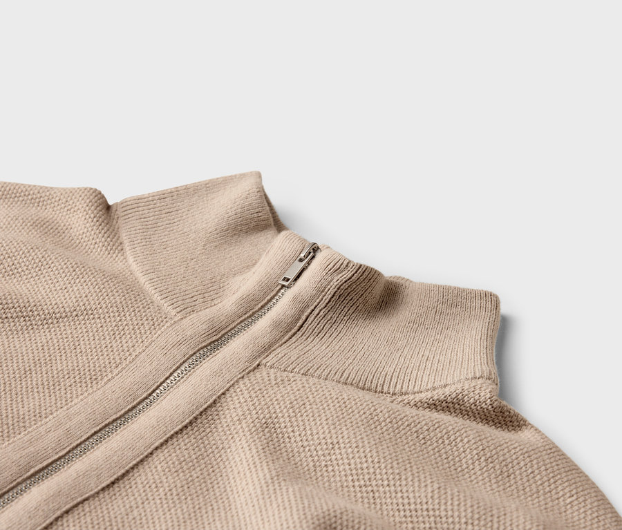 Nahaufnahme eines beigen Strickpullovers mit Reißverschluss und hohem Kragen.