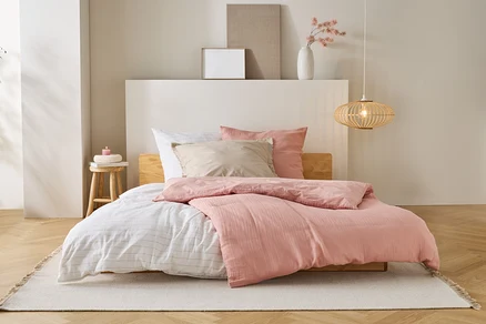 Ein Bett mit rosa Bettdecke, weißen Laken und beigen Kissen steht auf einem Teppich in einem Schlafzimmer.