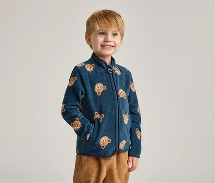 Ein lächelnder Junge mit blonden Haaren trägt eine marineblaue Fleecejacke mit Bärenmotiven und braune Cordhosen.