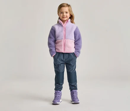 Mädchen mit lila-rosa Fleecejacke, blauen Hosen und lila Stiefeln steht vor hellem Hintergrund.