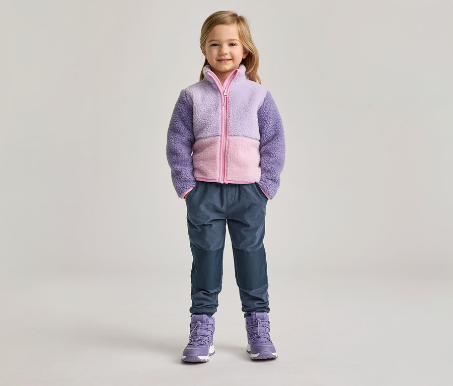 Mädchen mit lila-rosa Fleecejacke, blauen Hosen und lila Stiefeln steht vor hellem Hintergrund.