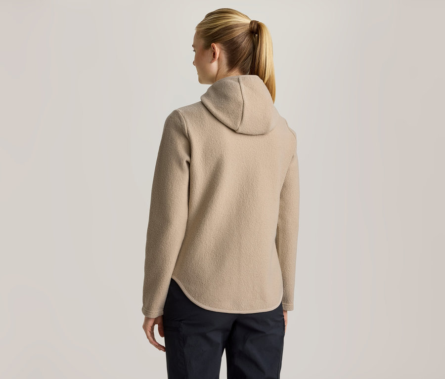 Frau von hinten, trägt eine beige Kapuzen-Fleecejacke und dunkle Hosen. Ihr blondes Haar ist zu einem Pferdeschwanz gebunden.