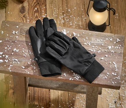 Ein Paar schwarze Handschuhe mit gerippten Bündchen und strukturierten Handflächen liegt auf einer rustikalen Holzbank, bedeckt mit Kunstschnee. Eine leuchtende Laterne und Tannenzapfen sind im Hintergrund auf dem Holzboden zu sehen.
