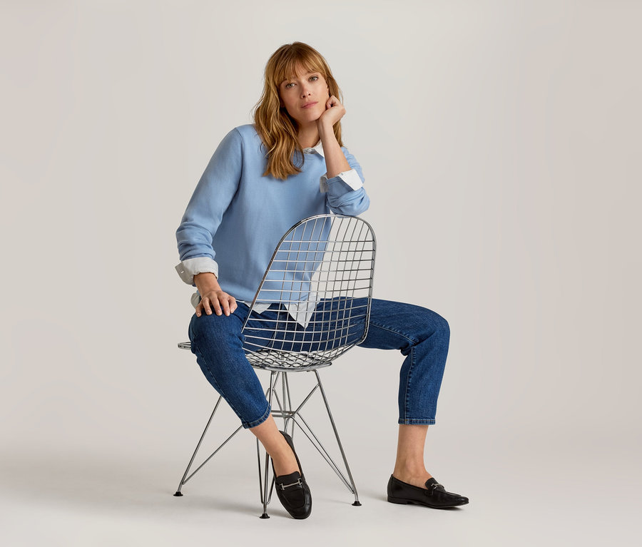 Frau mit hellbraunem Haar sitzt auf einem Metallstuhl, trägt ein hellblaues Sweatshirt, ein weißes Hemd, blaue Cropped-Jeans und schwarze Loafer, blickt in die Kamera.
