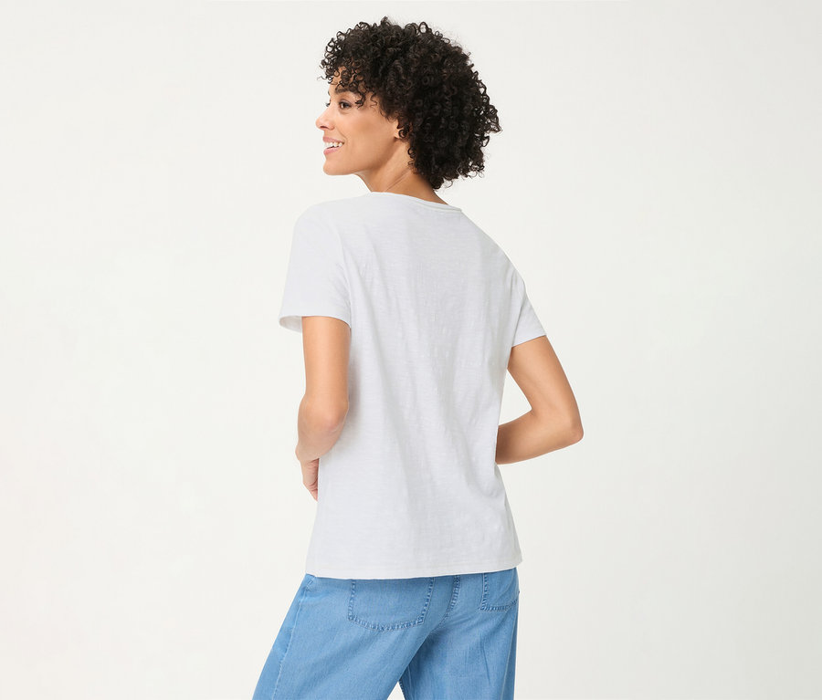 Frau mit lockigem Haar, von hinten gesehen, trägt ein weißes Olsen T-Shirt und hellblaue Hose, lächelt.
