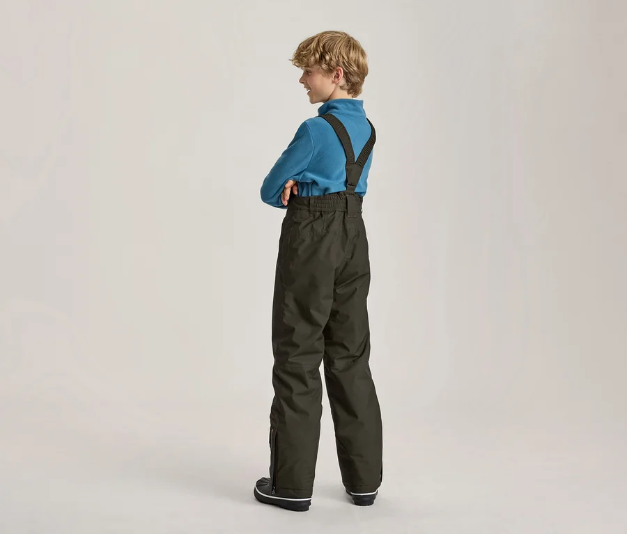 Junge mit blonden Locken, von hinten rechts gesehen, trägt ein blaues Langarmshirt und dunkelgrüne Skihose mit Hosenträgern, die Arme verschränkt und lächelt.