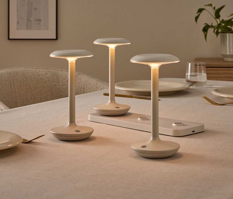 Drei minimalistische, cremefarbene Tischlampen auf einem gedeckten Esstisch, eine davon auf einer Ladestation. Die Lampen leuchten warm.