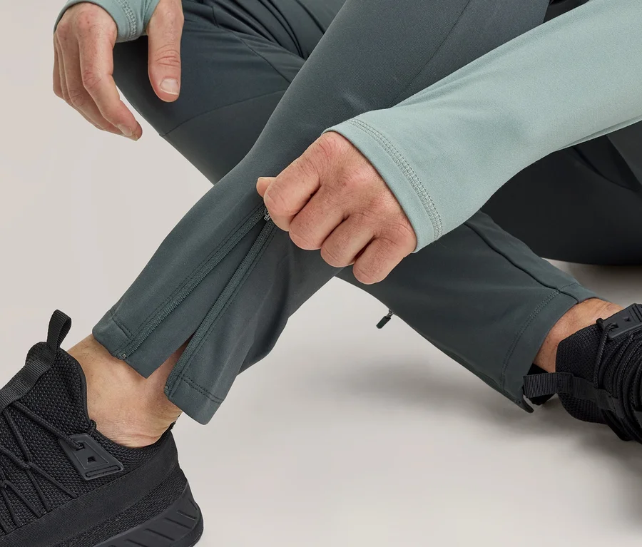 Eine Person in dunkelgrauer Sporthose und hellgrünem Oberteil schließt den Reißverschluss am Knöchel der Hose. Schwarze Sportschuhe sind sichtbar.