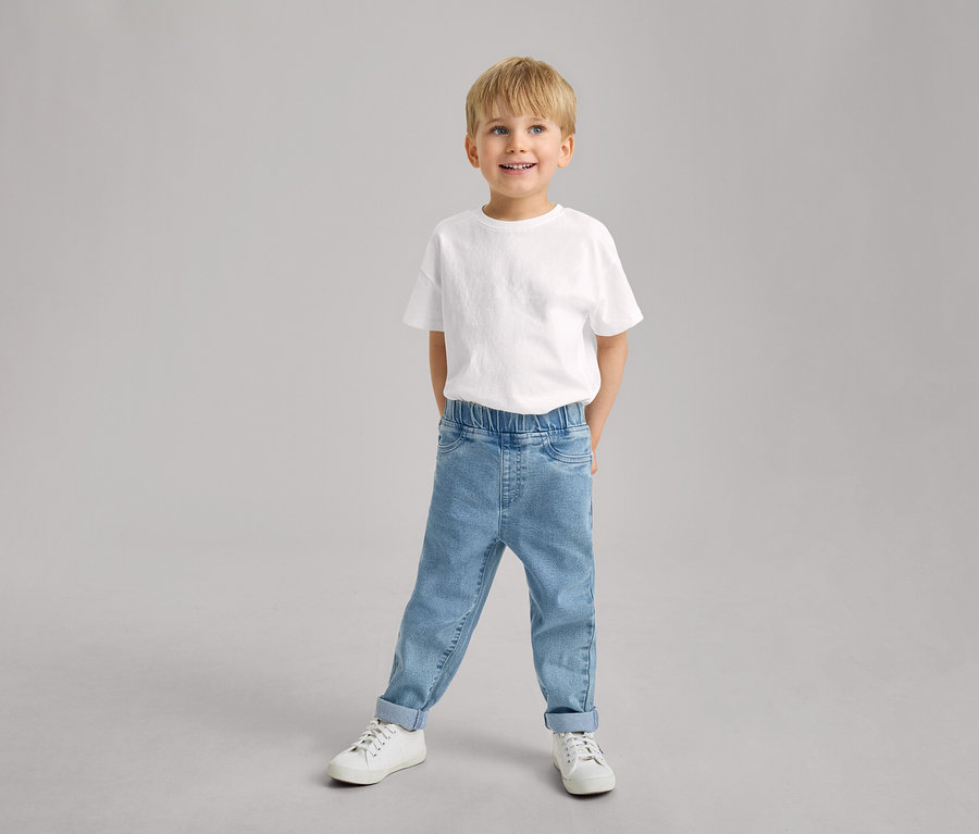 Ein lächelnder kleiner Junge steht in einem weißen T-Shirt, hellblauen Kinder-Pull-on-Pants – Fit »Luca« mit hochgekrempelten Hosenbeinen und weißen Turnschuhen.