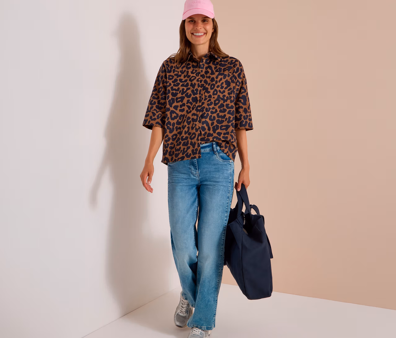 Lachende Frau mit rosa Kappe, hellblauer Jeans, silbernen Sneakers und einer braun-schwarz gemusterten CECIL Hemdbluse, eine dunkelblaue Tragetasche tragend.