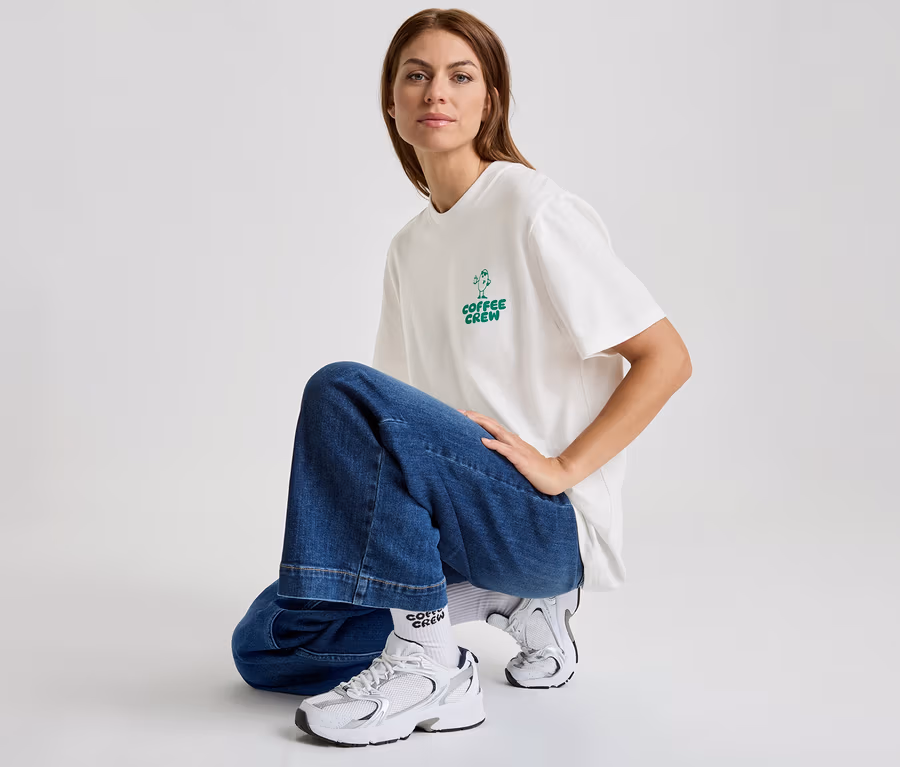 Frau hockt in Jeans und T-Shirt mit dem Aufdruck "Coffee Crew" und trägt weiße Turnschuhe.
