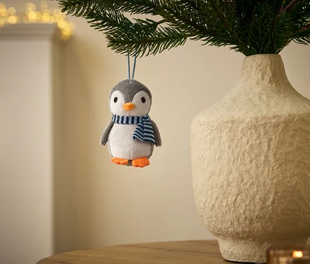 Ein grau-weißer Plüschpinguin-Anhänger mit orangefarbenem Schnabel und Füßen, der einen blau-weiß gestreiften Schal trägt, hängt an einem grünen Zweig. Daneben steht eine strukturierte, hellfarbene Vase mit Zweigen, im Hintergrund sind verschwommene Lichter zu sehen.
