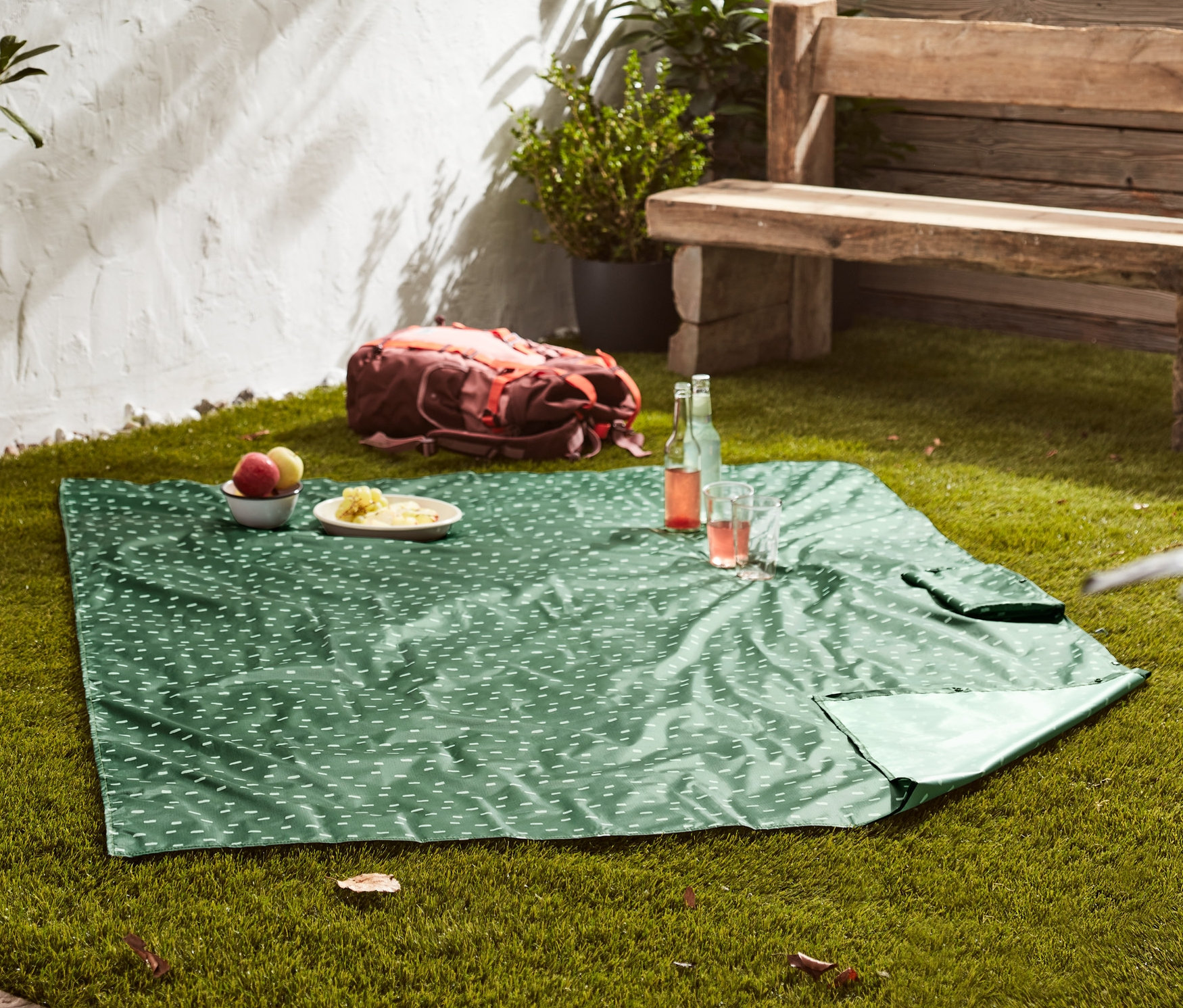 Eine grüne 2-in-1-Picknickdecke mit weissem Muster liegt auf einer Wiese. Auf der Decke stehen Teller mit Essen und Getränke.