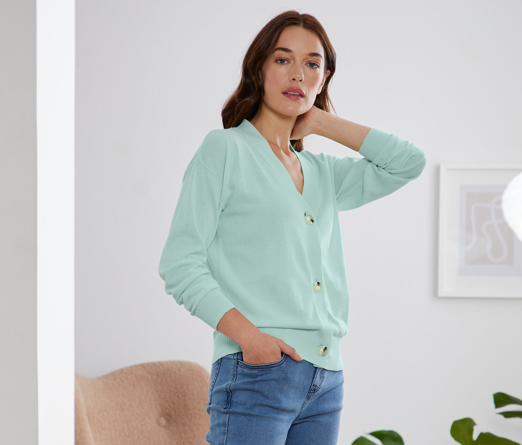 Frau posiert in mintfarbenem Cardigan mit V-Ausschnitt, Jeans und weißen Turnschuhen.