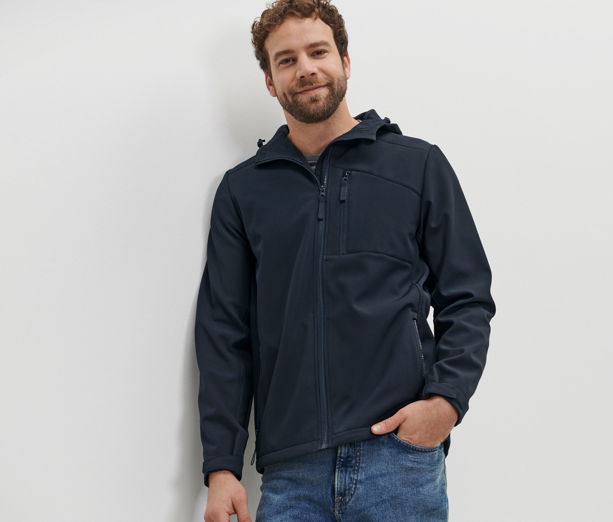 Mann lehnt an Wand, trägt Softshelljacke.