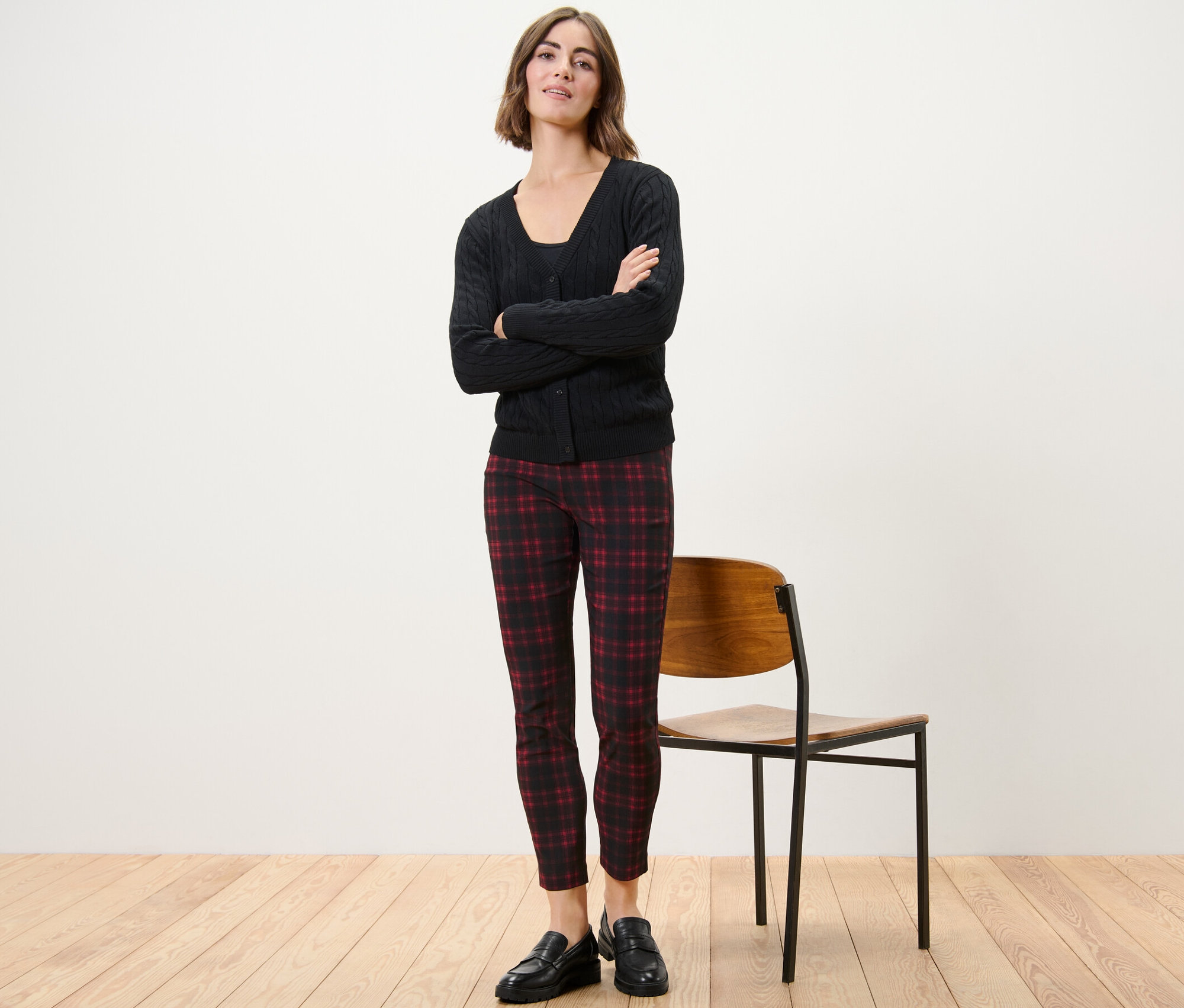 Eine Frau steht mit verschränkten Armen und trägt eine karierte Stretchhose, eine schwarze Jacke und schwarze Schuhe.