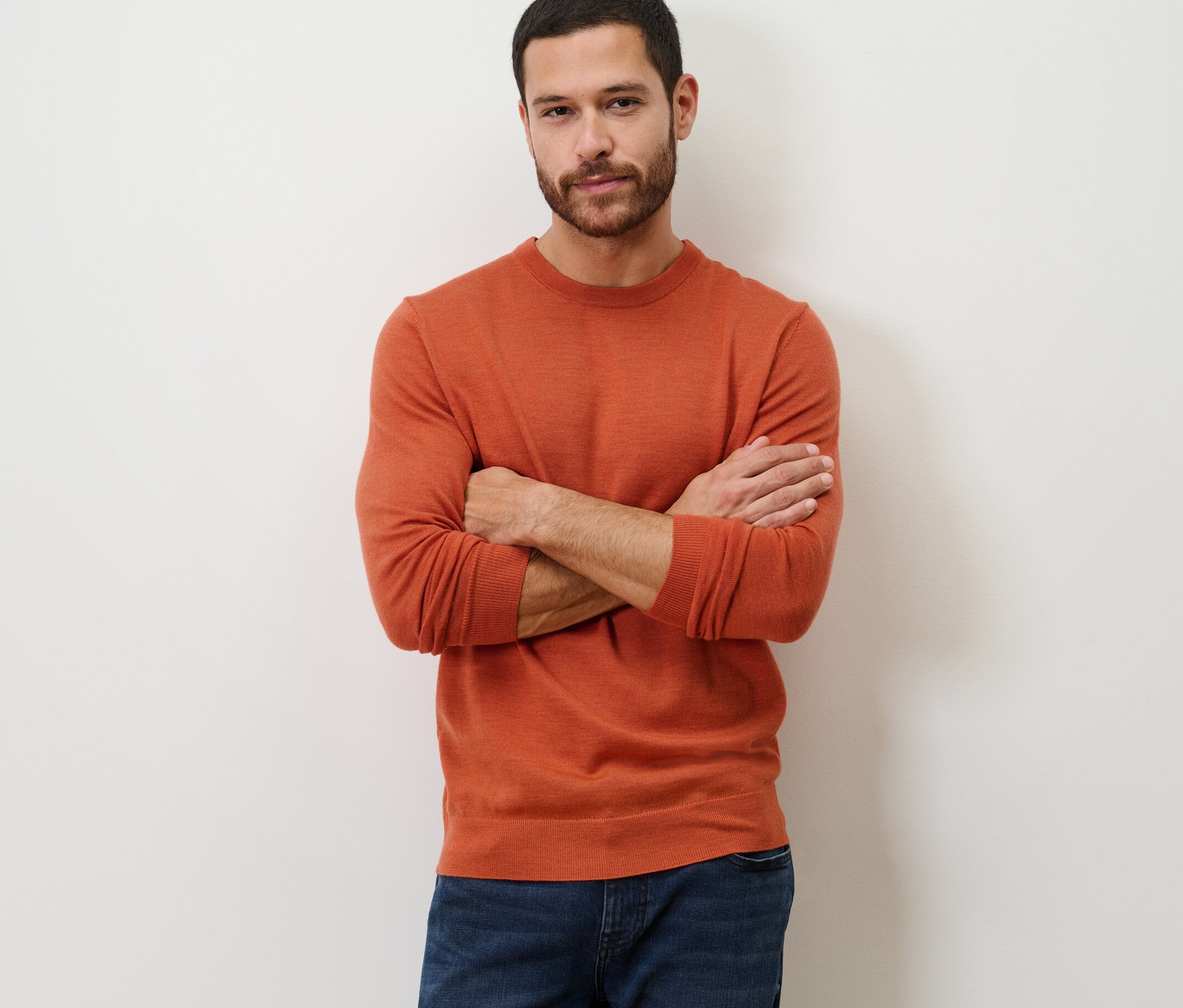 Mann mit verschränkten Armen posiert in orangefarbenem Merino-Pullover.