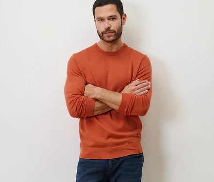 Mann mit verschränkten Armen posiert in orangefarbenem Merino-Pullover.