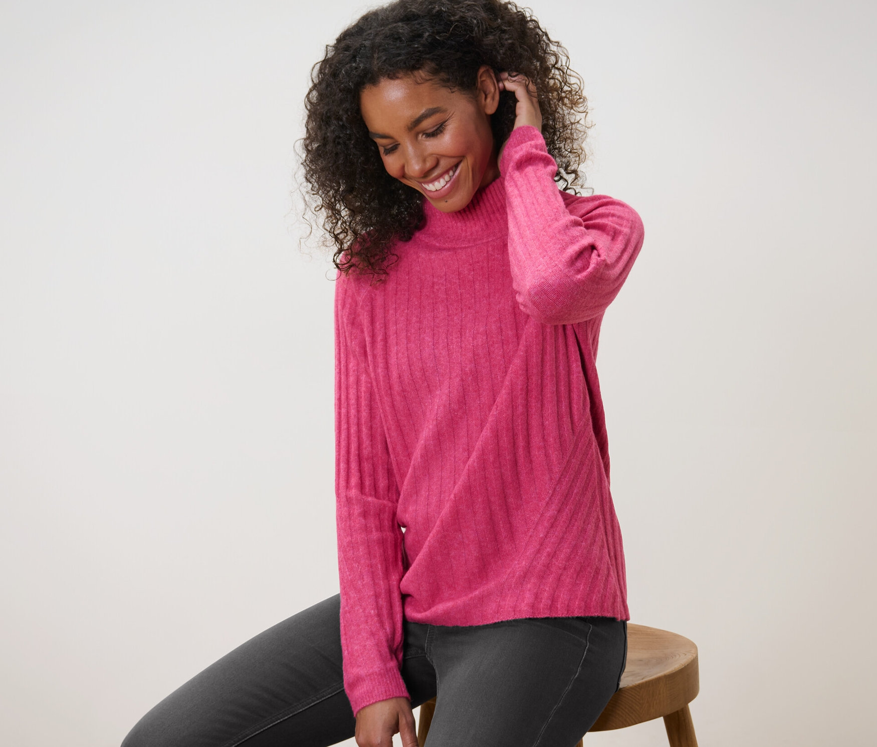 Frau sitzt auf einem Hocker und trägt einen gerippten Strickpullover mit Stehkragen.