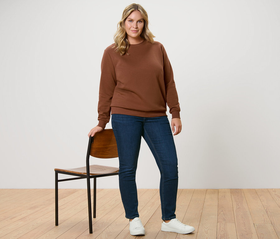 Eine Frau steht neben einem Stuhl und trägt ein braunes Sweatshirt und eine Slim Jeans – Fit »Emma« in Dunkelblau.
